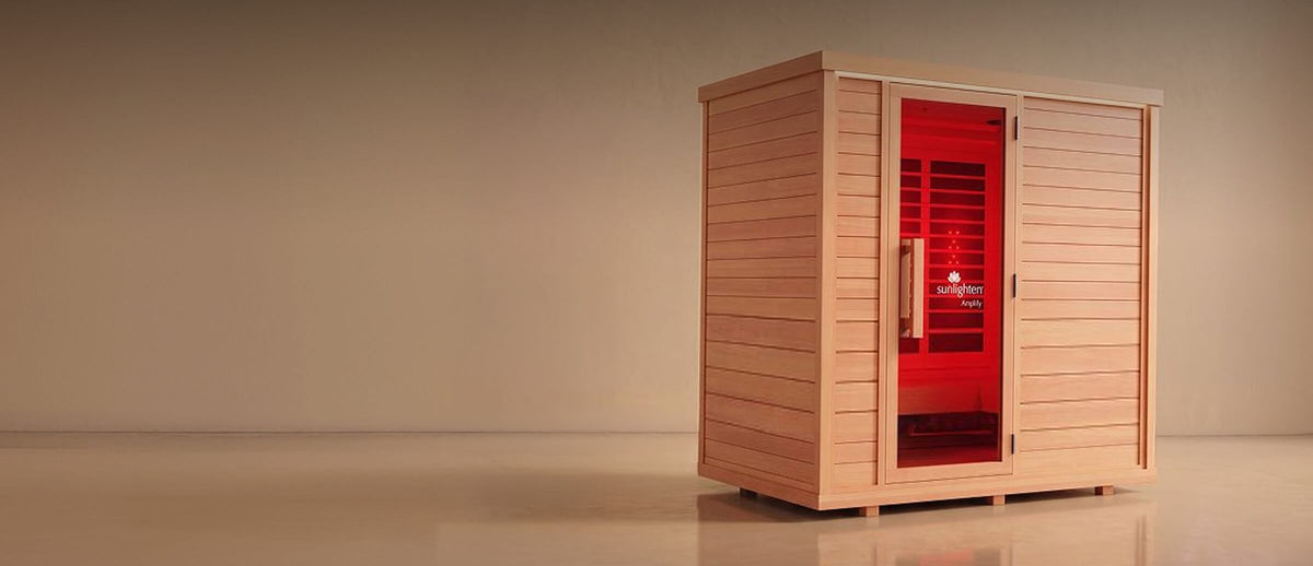 A wooden infrared sauna cabin with a glass door showing a red interior, set against a plain beige studio background.