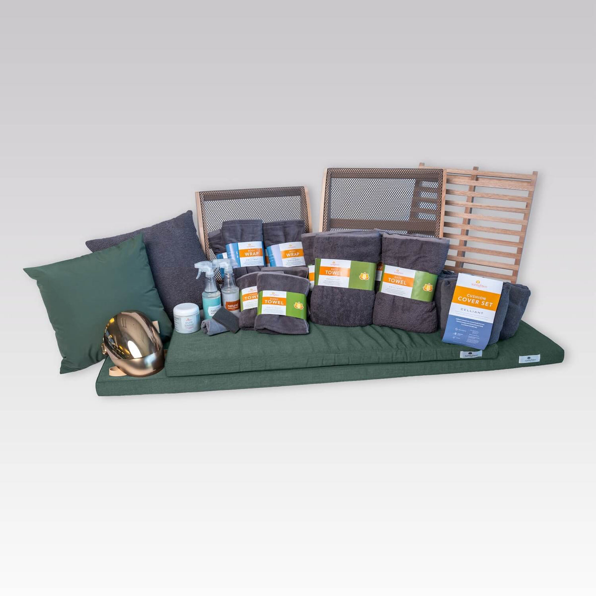 A collection of dark gray towels and accessories arranged on a light gray background, including a large throw pillow, several folded towels, a cleaning kit, a jar of Pure Sweat cream, a gold RED Light Therapy Mask, and two wooden sauna backrests.