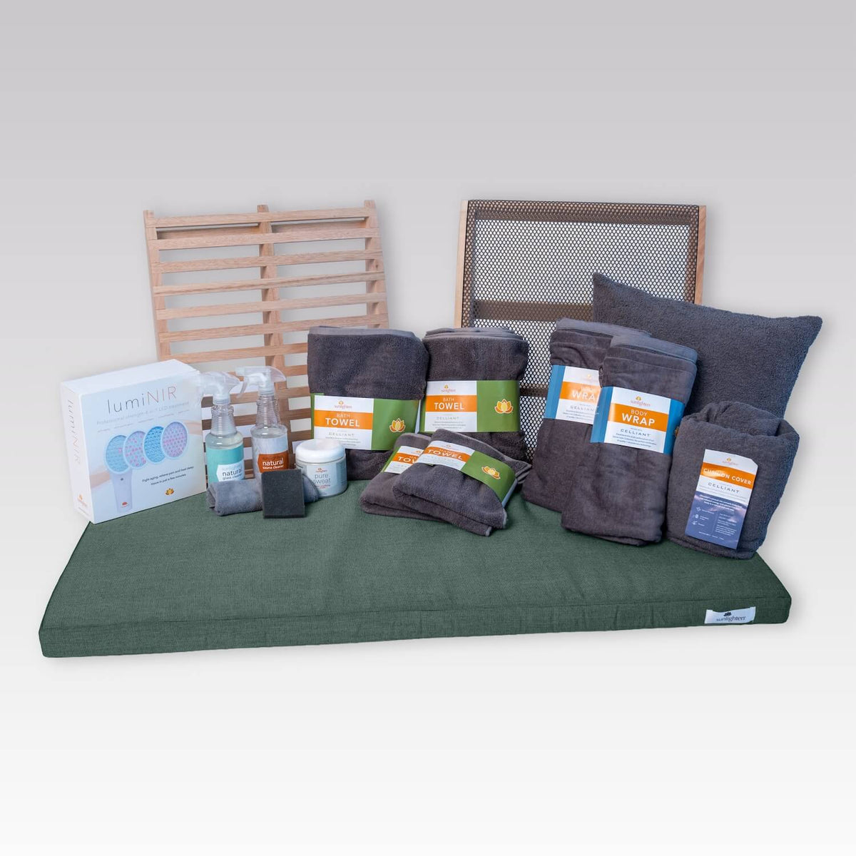 A collection of dark gray towels and accessories arranged on a light gray background, including a large throw pillow, several folded towels, a cleaning kit, a jar of Pure Sweat cream, a LumiNIR box, and wooden sauna backrests.