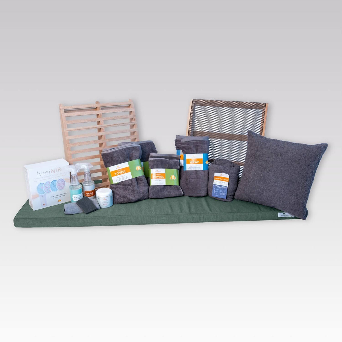 A sauna bench cushion set displayed on a white studio background, featuring a cushion, matching green and dark gray pillows, dark gray towels, packaged cleaning supplies, and wooden backrests.