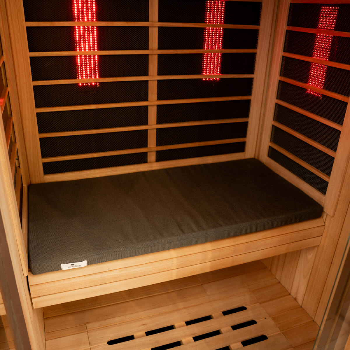 A wooden infrared sauna room with a dark gray cushioned bench, black mesh walls, and glowing red LED light panels.