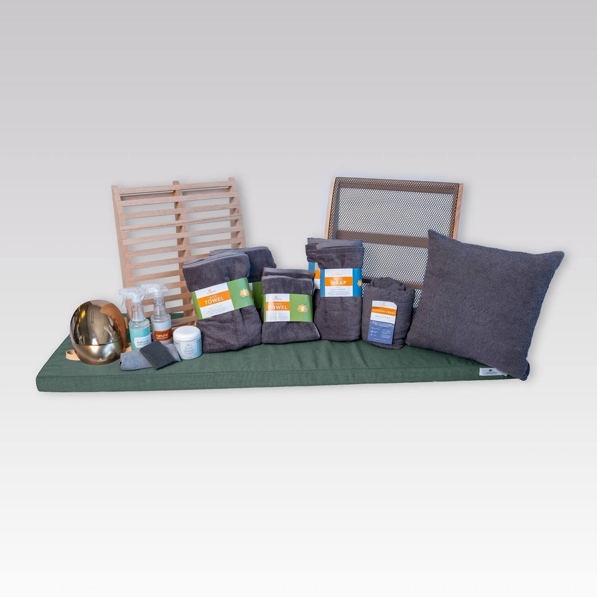A collection of dark gray towels and accessories arranged on a light gray background, including a large throw pillow, several folded towels, a cleaning kit, a jar of Pure Sweat cream, a gold RED Light Therapy Mask, and two wooden sauna backrests.