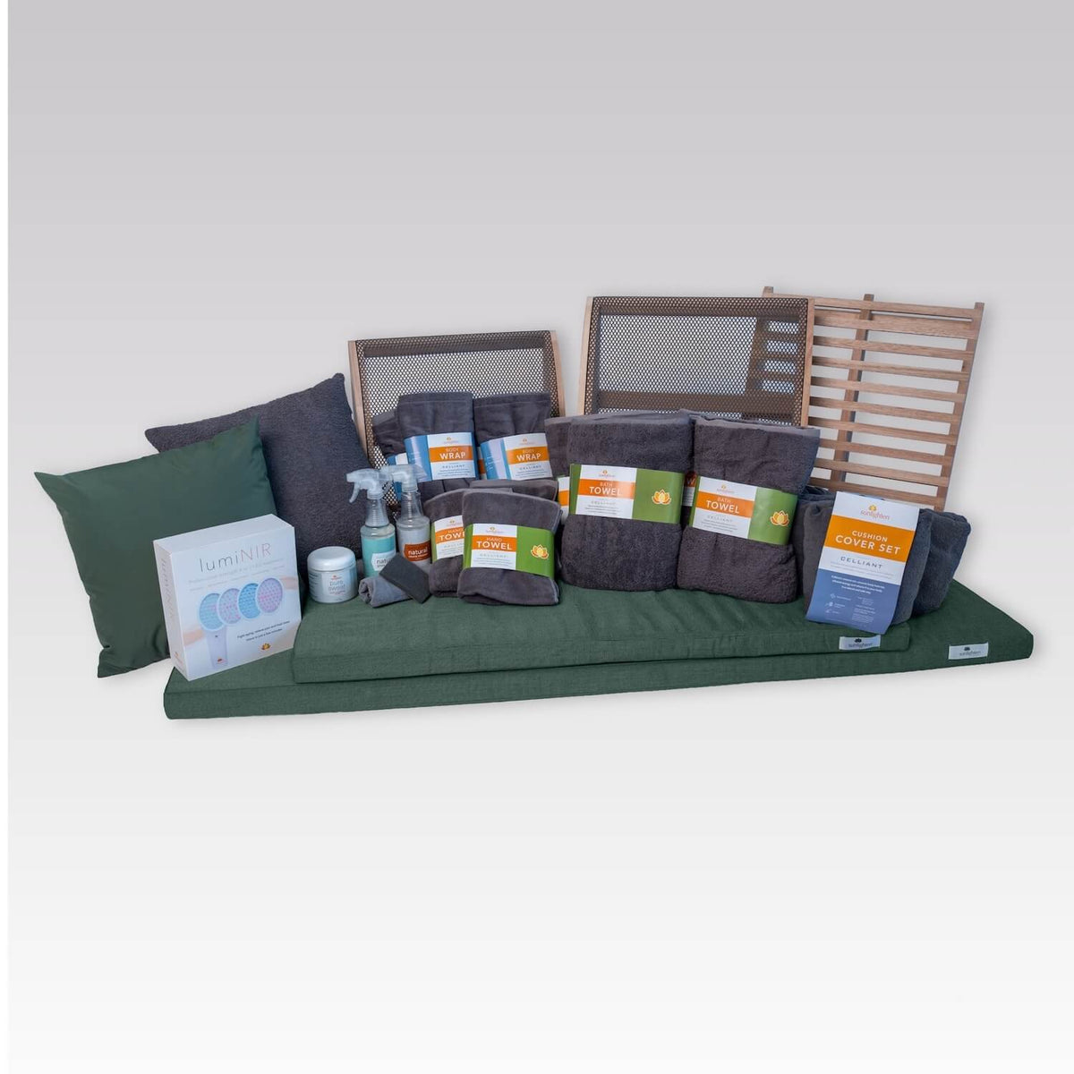 A sauna bench cushion set displayed on a white studio background, featuring a cushion, matching green and dark gray pillows, dark gray towels, packaged cleaning supplies, and wooden backrests.