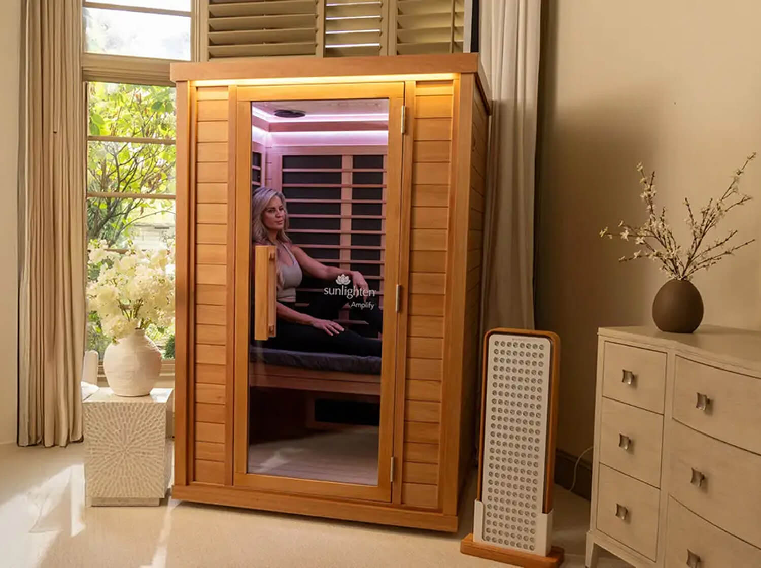 A AMPLIFY infrared sauna cabin with a glass door, shown in a styled room with a woman sitting inside. A portable sauna pad holder stands next to it on the floor.