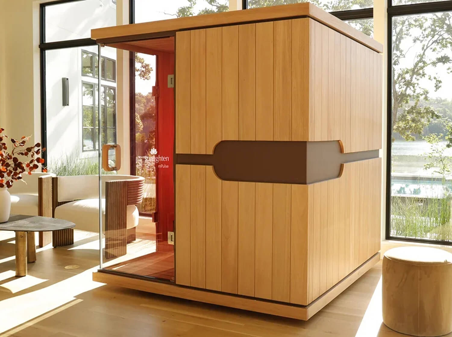 An mPulse wooden sauna cabin with a light natural wood exterior, a dark brown accent band, and a glass door, shown in a bright contemporary room with large windows.