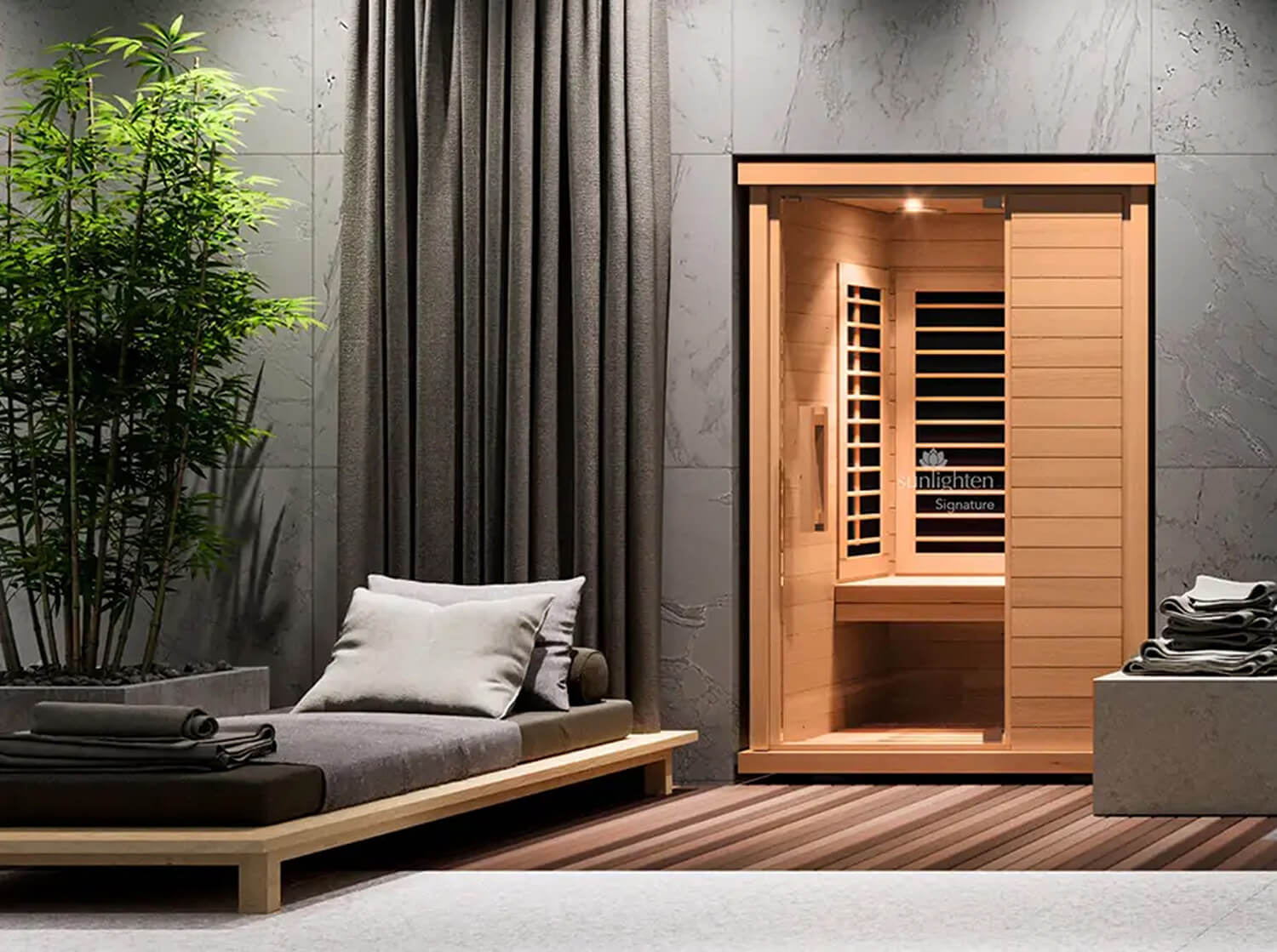 A SUNLIGHTEN SIGNATURE wooden indoor sauna with sliding doors stands in a styled room with gray stone walls, dark curtains, a low wooden daybed with cushions, and a potted bamboo plant.