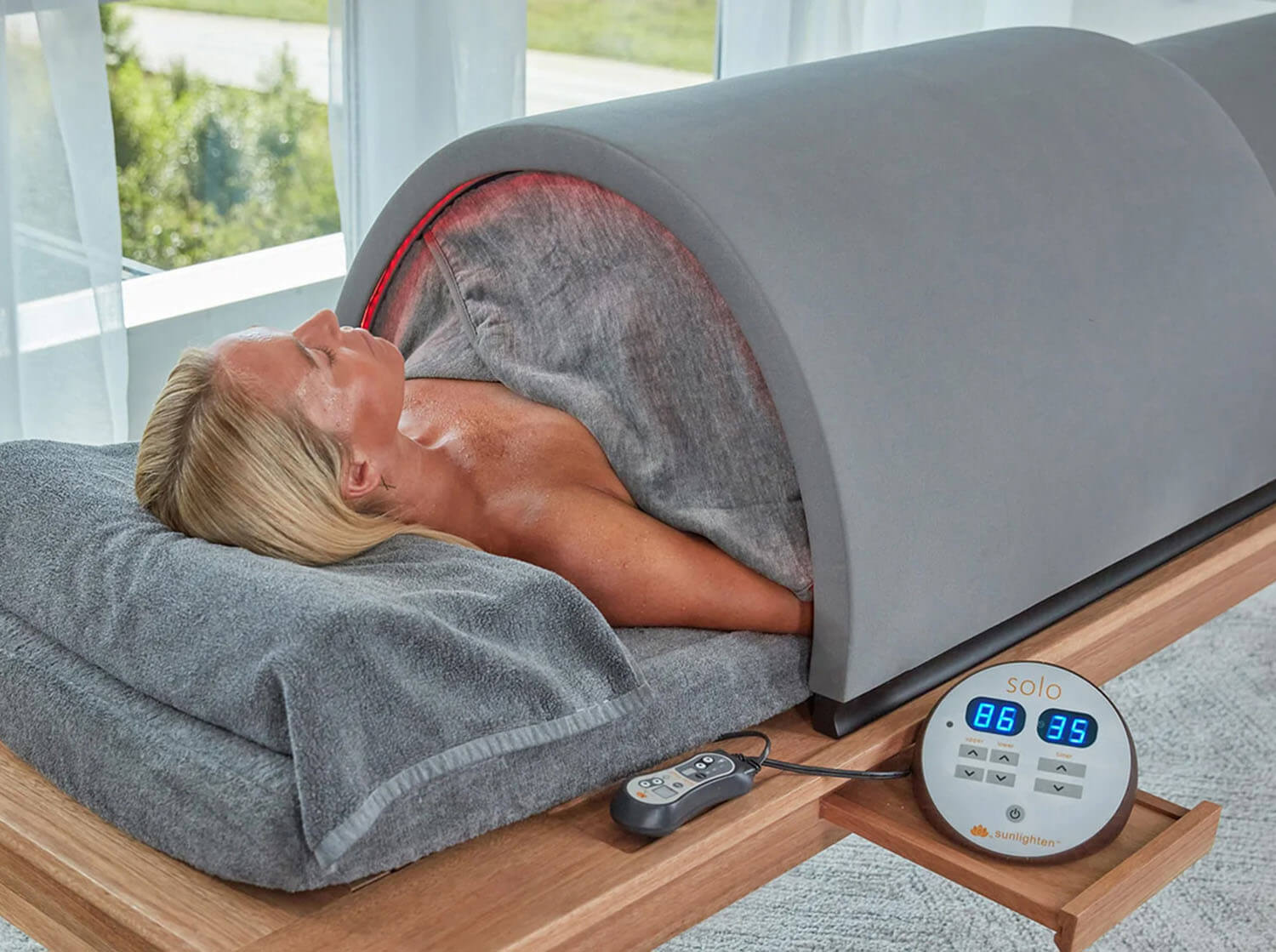 A person lying on a gray Solo infrared sauna body treatment bed with a pillow and blanket, shown in a bright indoor room. The bed has a digital control panel on a wooden stand.